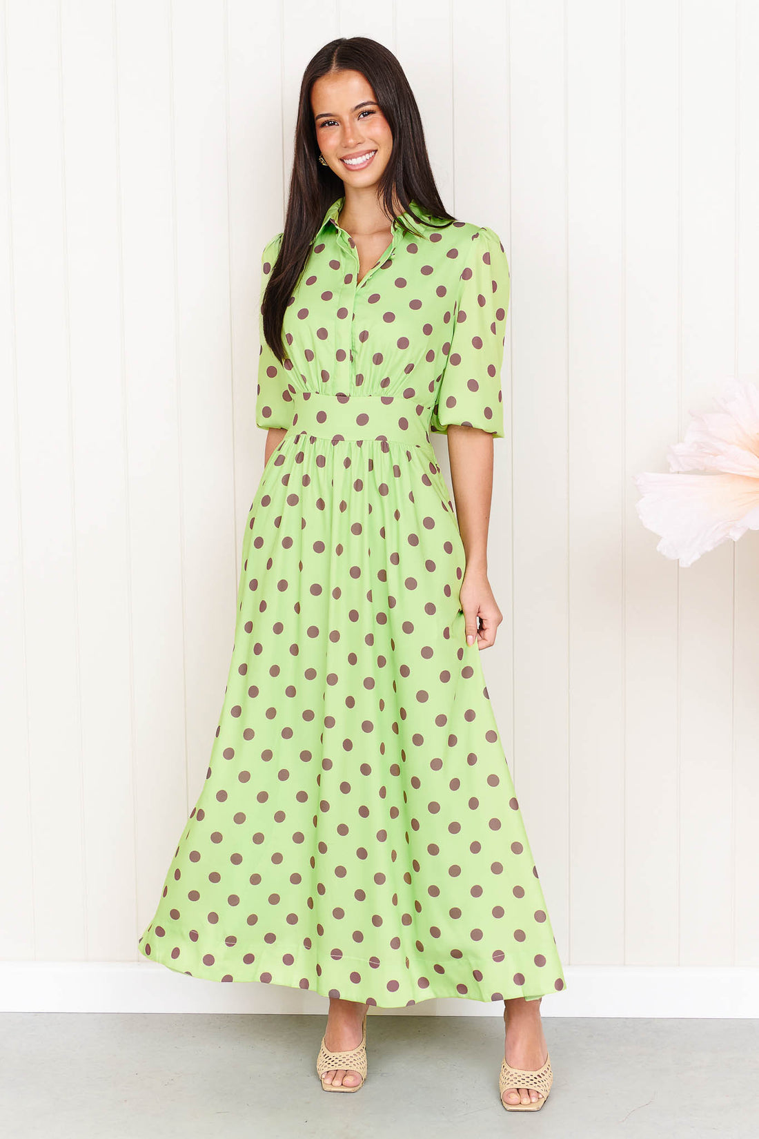 A woman wearing the minnie + MAXXIE Dolce Midi event dress in lime with chocolate brown polka dots, standing in front of a white panelled wall.