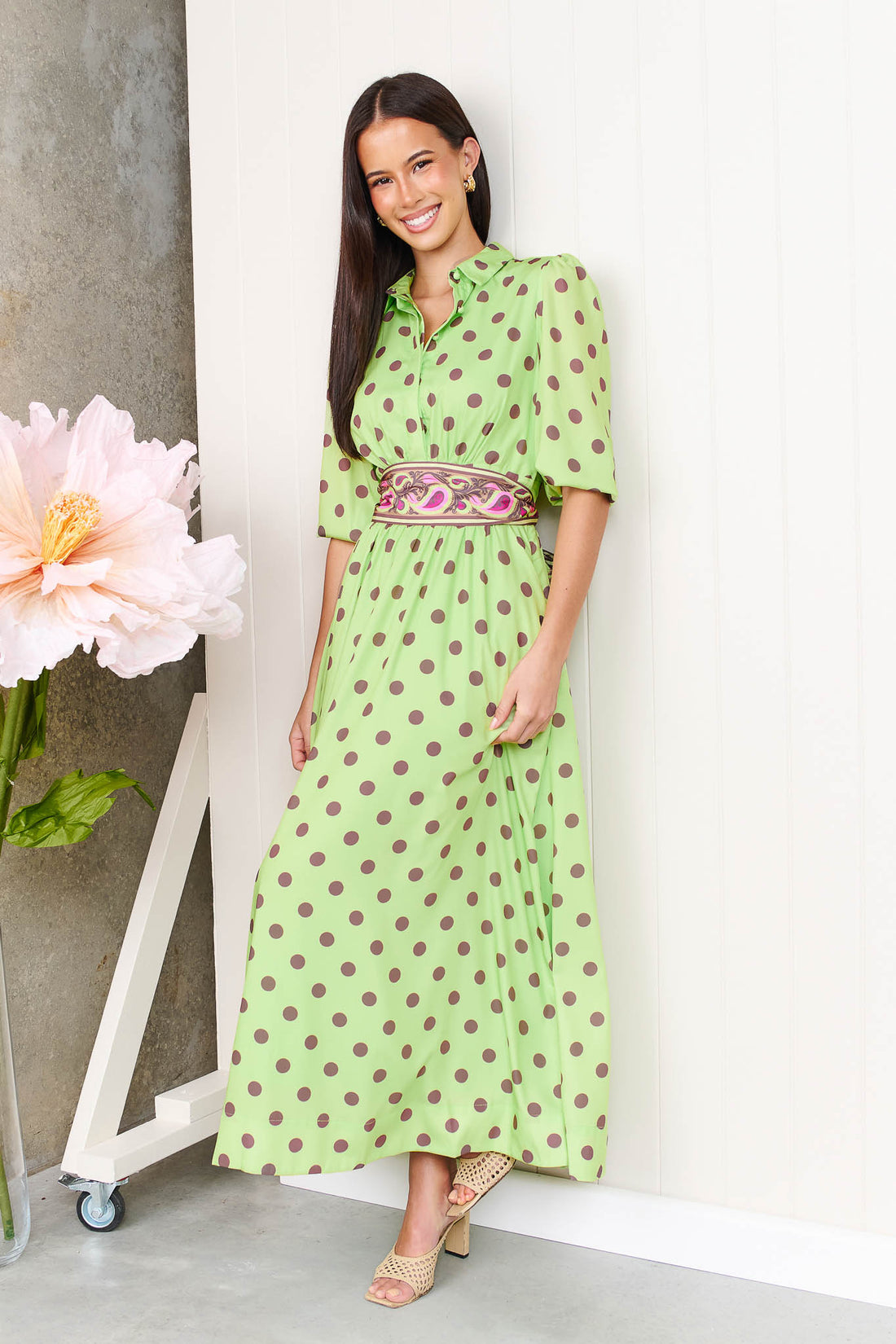 A woman wearing the minnie + MAXXIE Dolce Midi event dress in lime with chocolate brown polka dots and a matching paisley patterned sash waist tie, standing in front of a white panelled wall.