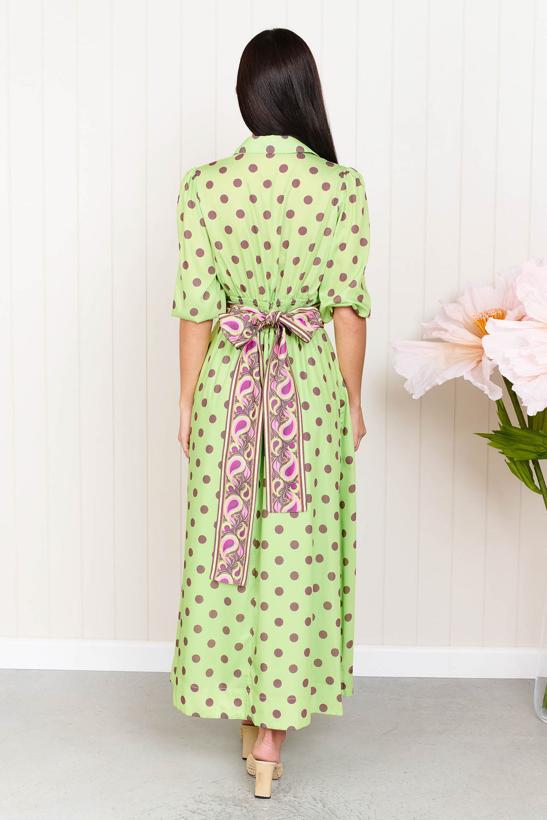 A woman wearing the minnie + MAXXIE Dolce Midi event dress in lime with chocolate brown polka dots and a matching paisley patterned sash waist tie, standing in front of a white panelled wall.