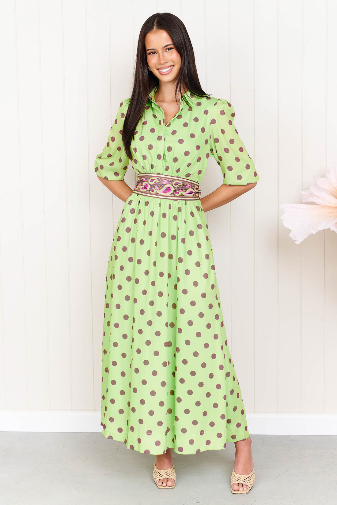 A woman wearing the minnie + MAXXIE Dolce Midi event dress in lime with chocolate brown polka dots and a matching paisley patterned sash waist tie, standing in front of a white panelled wall.