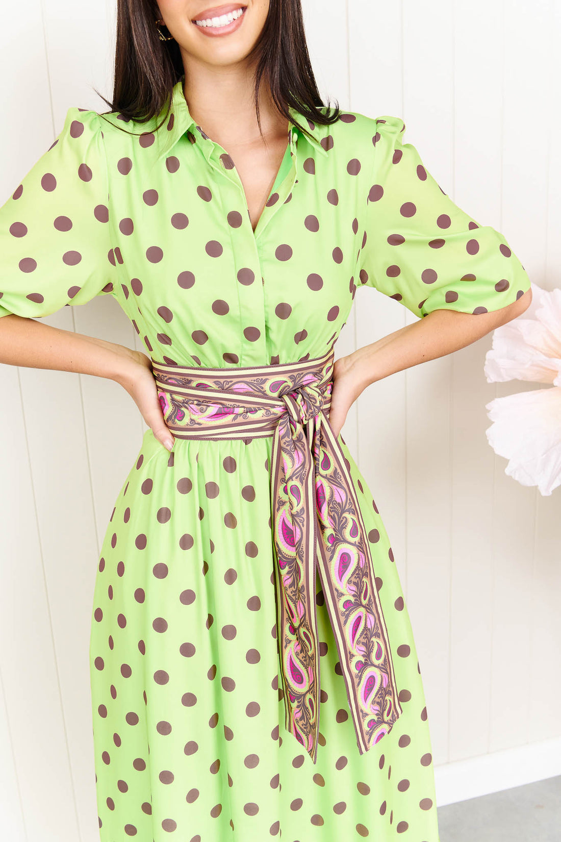 A woman wearing the minnie + MAXXIE Dolce Midi event dress in lime with chocolate brown polka dots and a matching paisley patterned sash waist tie, standing in front of a white panelled wall.