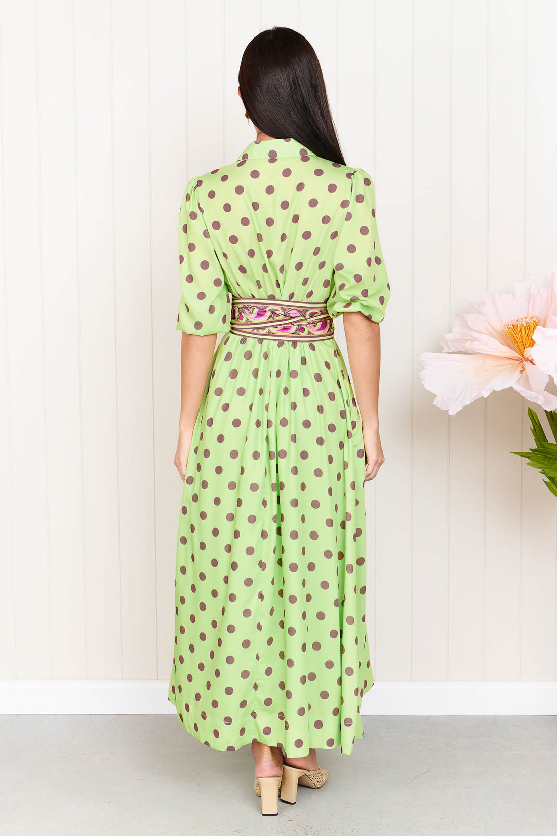 Back view of a woman wearing the minnie + MAXXIE Dolce Midi event dress in lime with chocolate brown polka dots and a matching paisley patterned sash waist tie, standing in front of a white panelled wall.