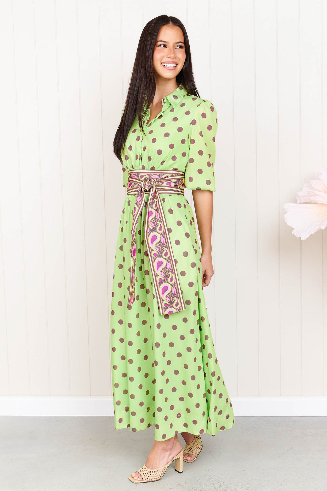 A woman wearing the minnie + MAXXIE Dolce Midi event dress in lime with chocolate brown polka dots and a matching paisley patterned sash waist tie, standing in front of a white panelled wall.
