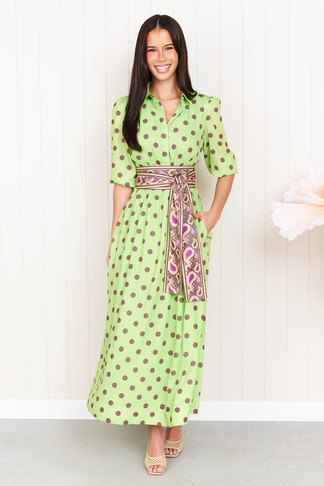 A woman wearing the minnie + MAXXIE Dolce Midi event dress in lime with chocolate brown polka dots and a matching paisley patterned sash waist tie, standing in front of a white panelled wall.