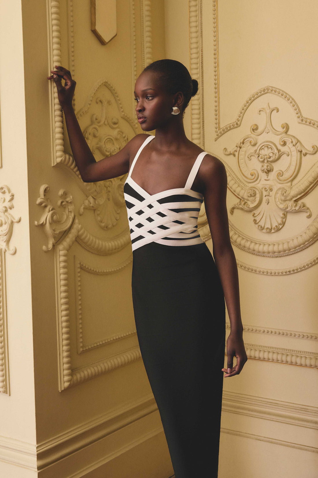 A model wearing the ELLIATT Ninette Column Midi Dress in Black & White with a white criss-cross strap pattern across the bodice, photographed against an opulent interior wall background.