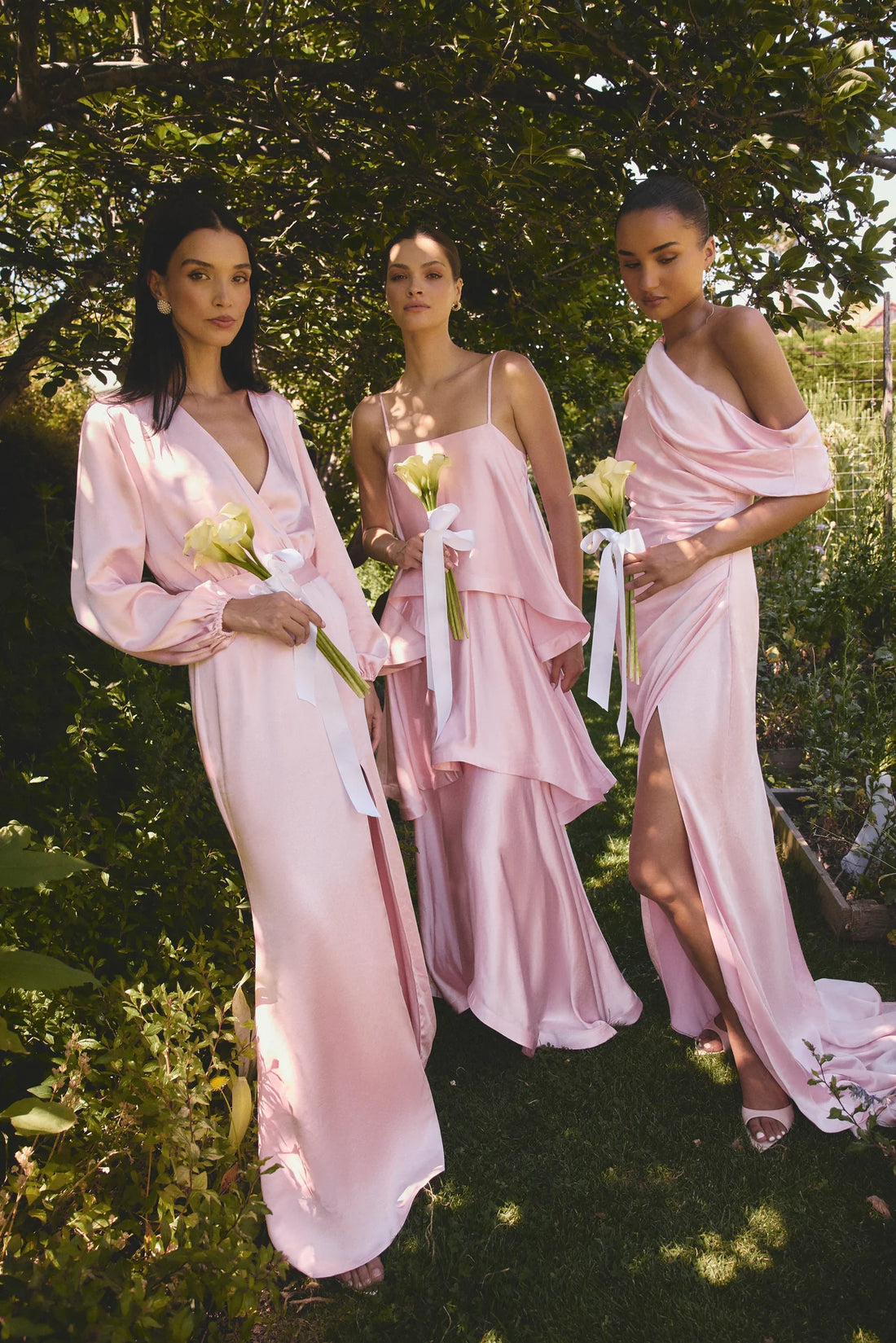Three bridesmaids in pink ELLIATT dresses standing outdoors with greenery around them.