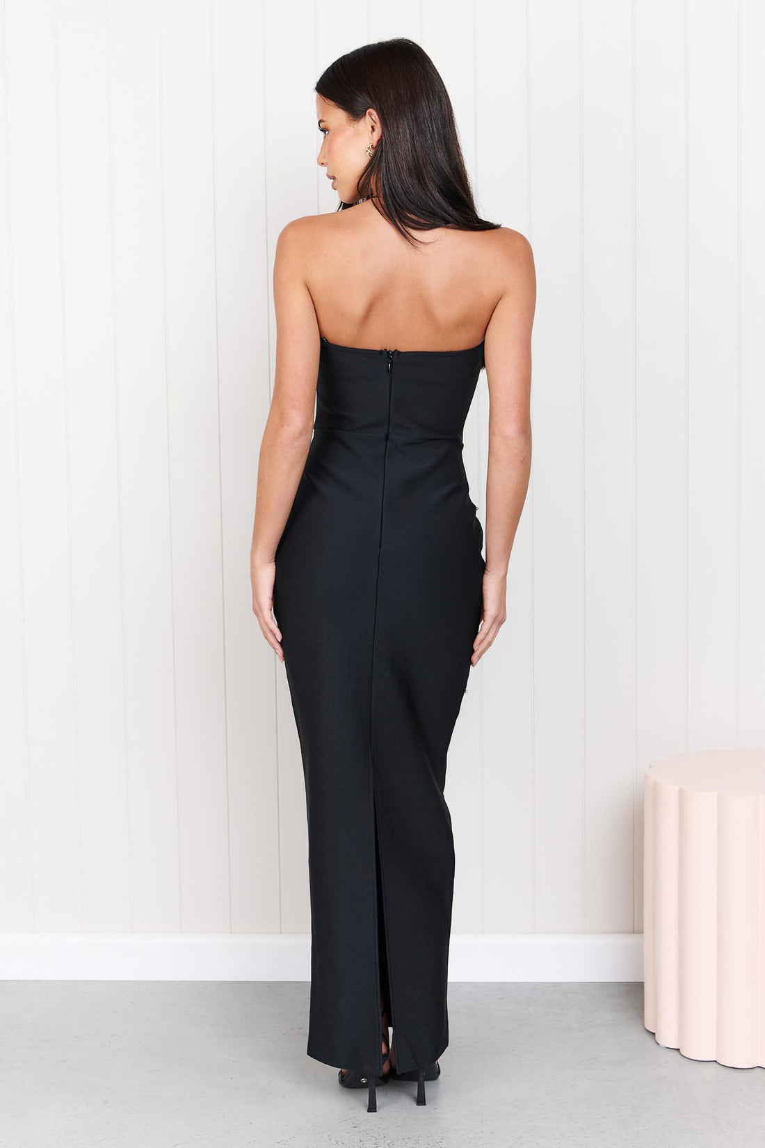 Back view of a woman modelling the ELLIATT Natasia embellished strapless black maxi dress with white floral appliqué flowers and pearl stud embellishments, photographed against a white panelled wall.