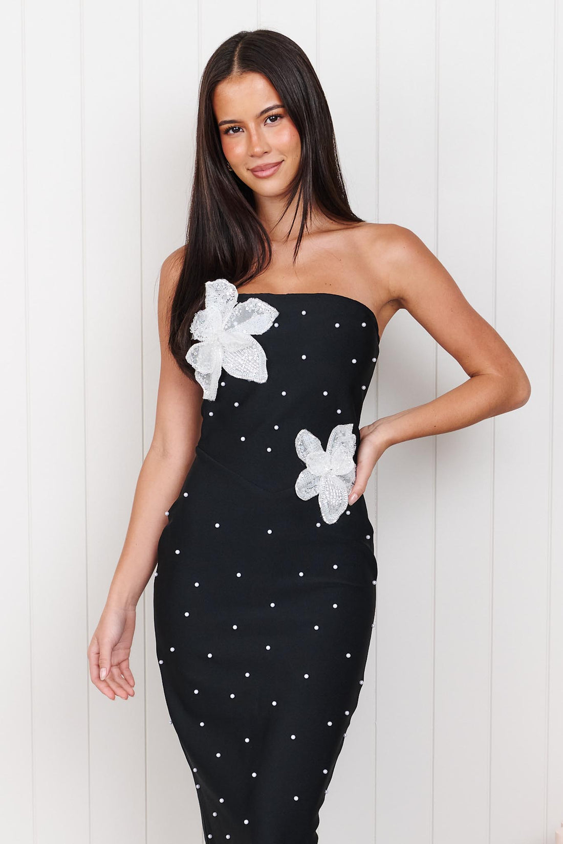 A woman modelling the ELLIATT Natasia embellished strapless black maxi dress with white floral appliqué flowers and pearl stud embellishments, photographed against a white panelled wall.