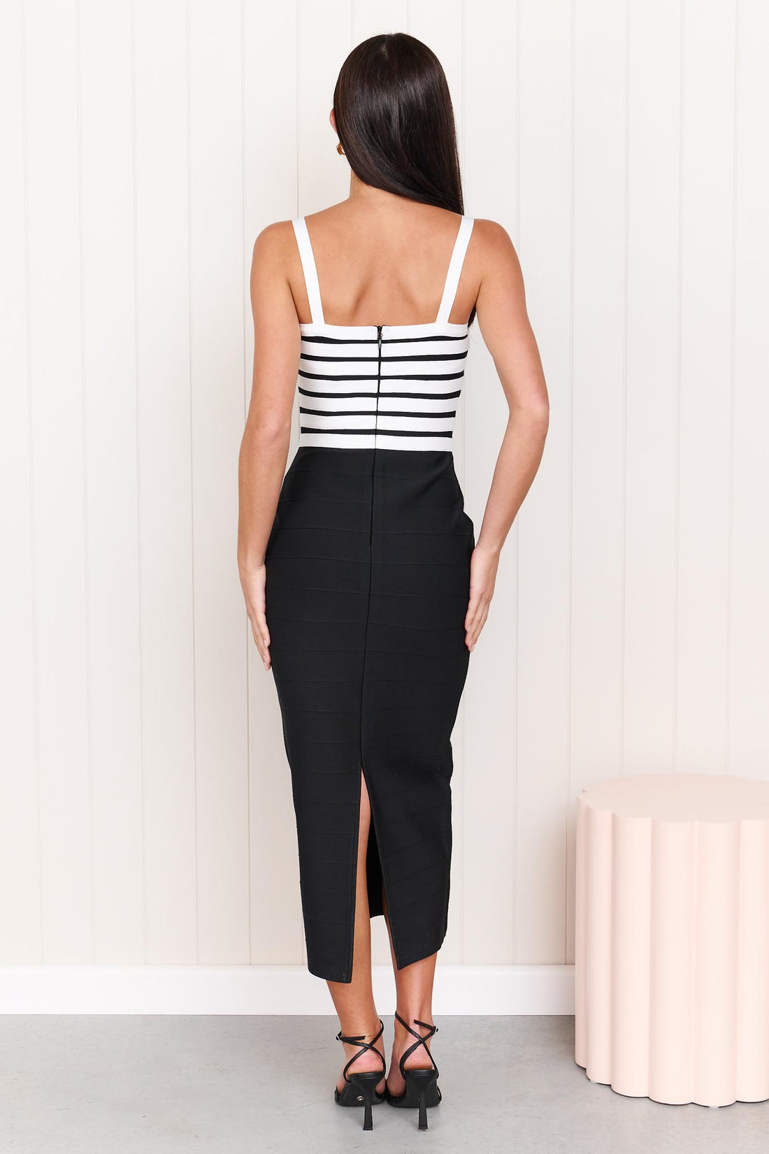 Back view of a model wearing the ELLIATT Ninette Column Midi Dress in Black & White with a white criss-cross strap pattern across the bodice, photographed against a white panelled wall.
