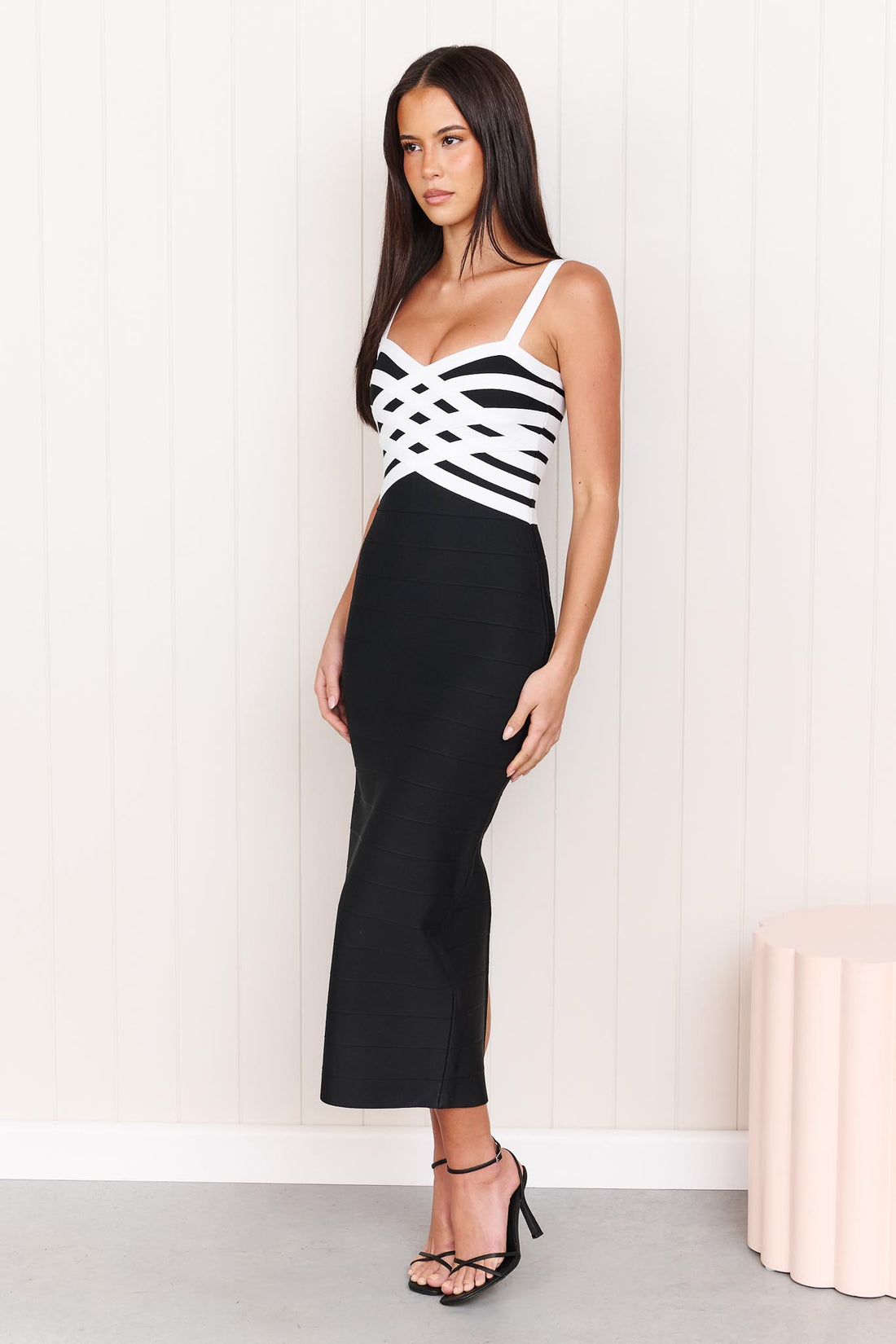 A model wearing the ELLIATT Ninette Column Midi Dress in Black & White with a white criss-cross strap pattern across the bodice, photographed against a white panelled wall.