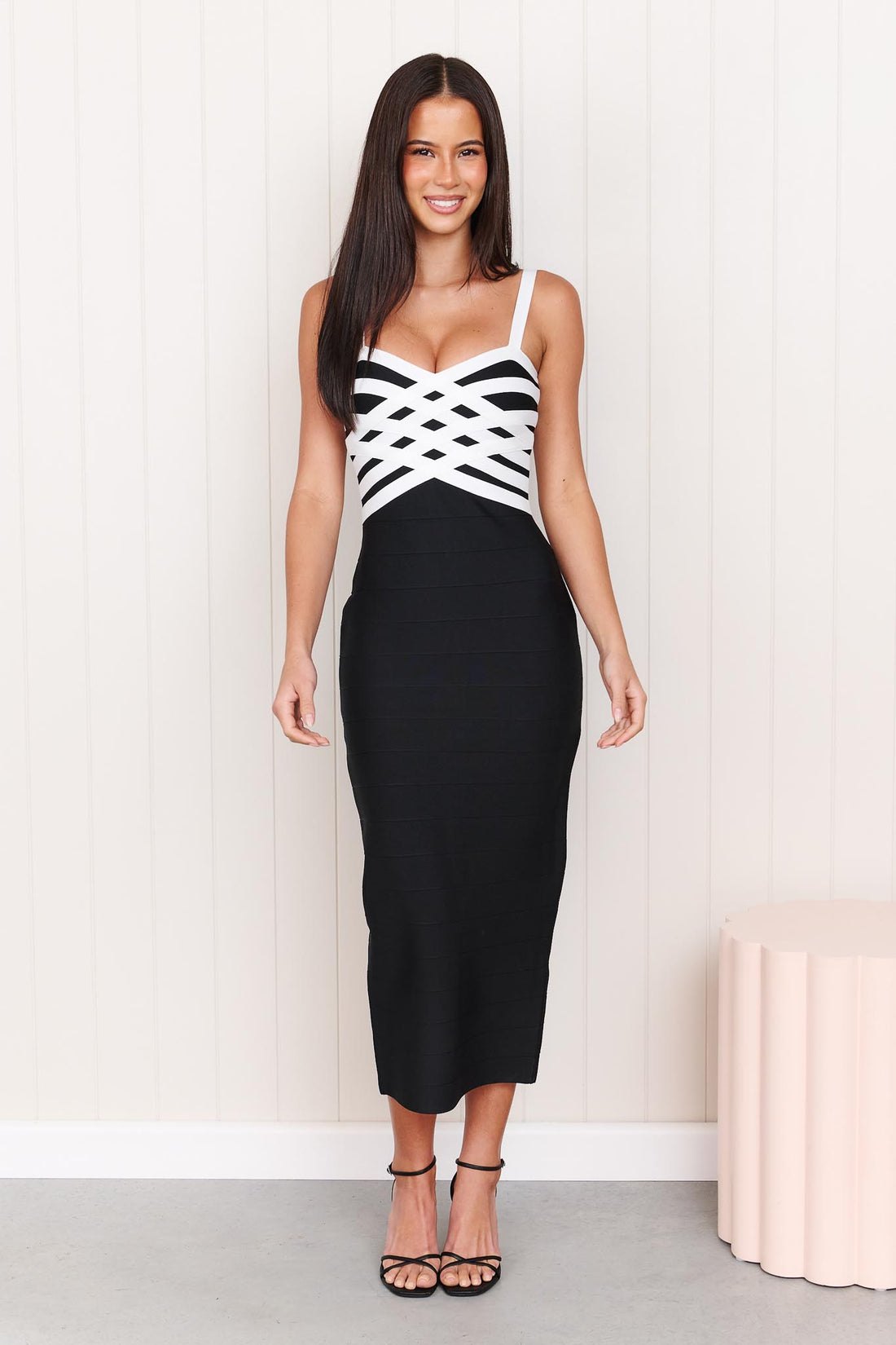 A model wearing the ELLIATT Ninette Column Midi Dress in Black & White with a white criss-cross strap pattern across the bodice, photographed against a white panelled wall.