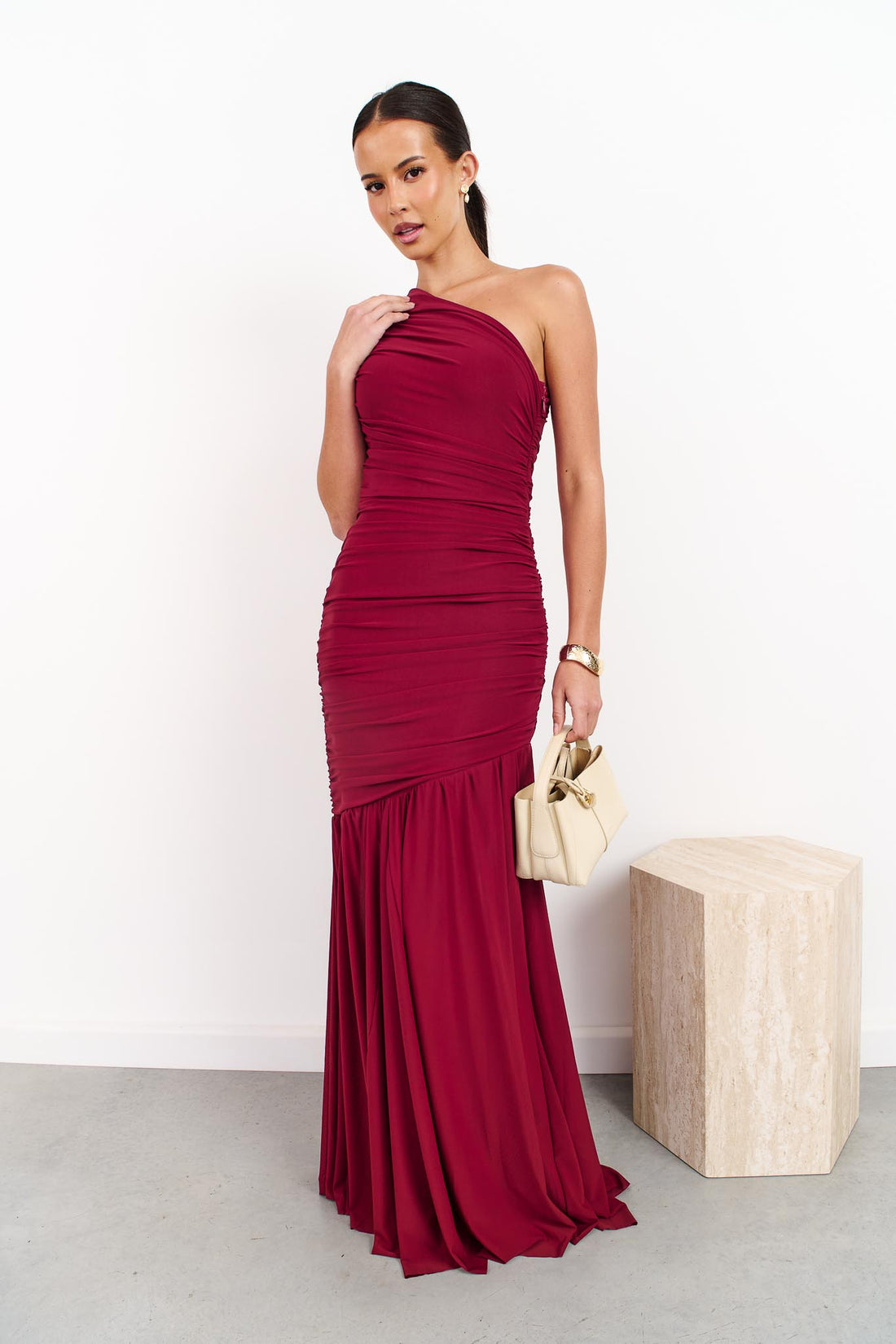 A woman wearing the ELLIATT Florencea one shoulder event gown in Wine Red, featuring a ruched bodice and draped maxi length skirt. She is standing in a front of a white wall.