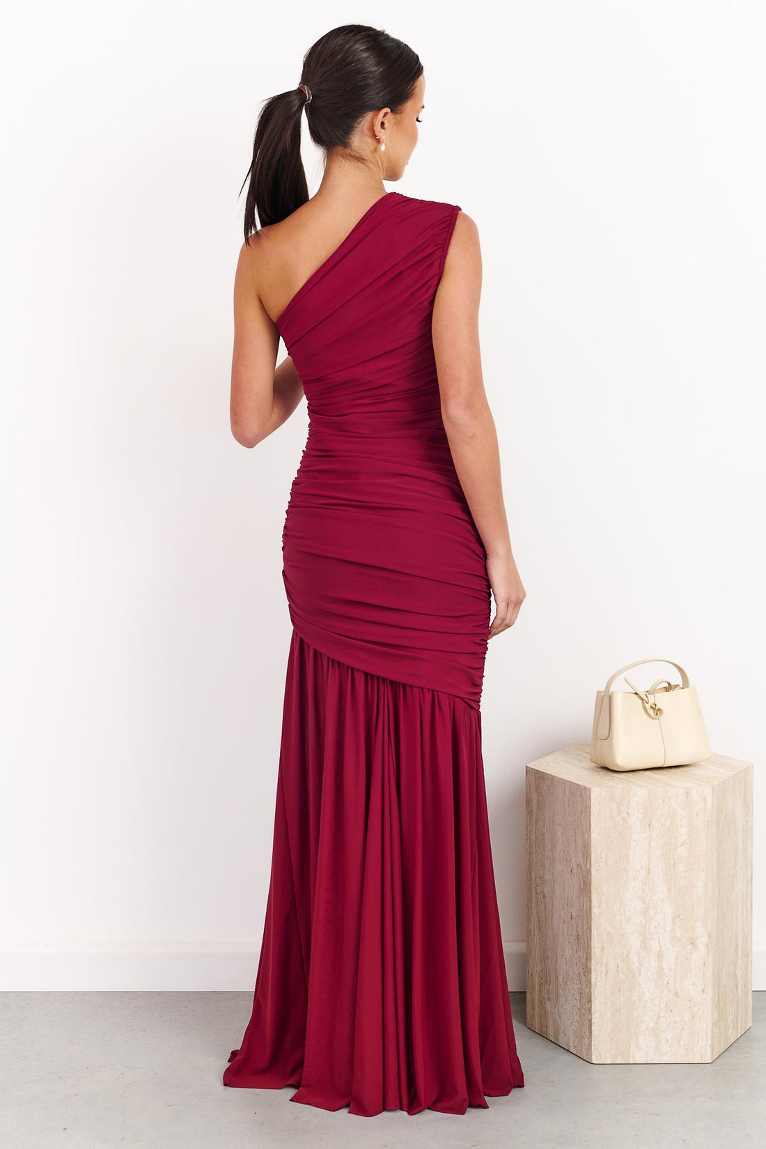 Back view of a woman wearing the ELLIATT Florencea one shoulder event gown in Wine Red, featuring a ruched bodice and draped maxi length skirt. She is standing in a front of a white wall.