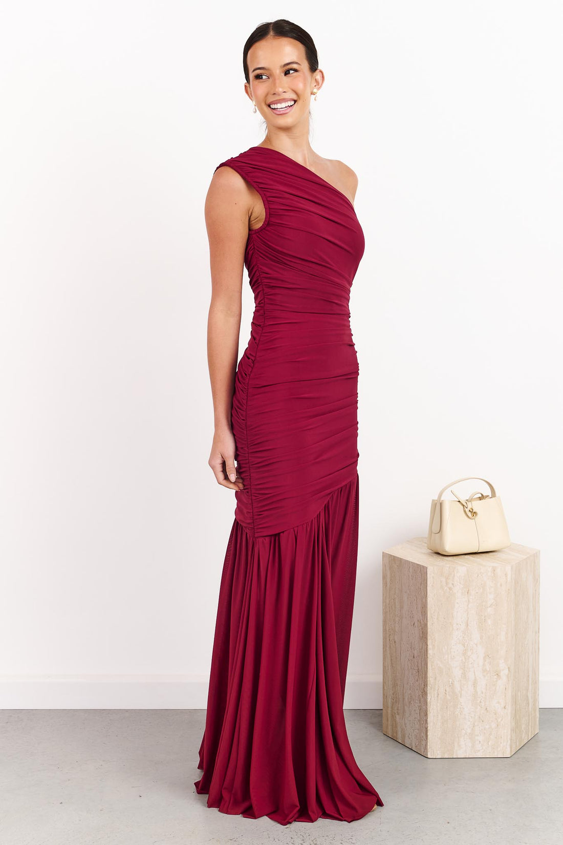 A woman wearing the ELLIATT Florencea one shoulder event gown in Wine Red, featuring a ruched bodice and draped maxi length skirt. She is standing in a front of a white wall.