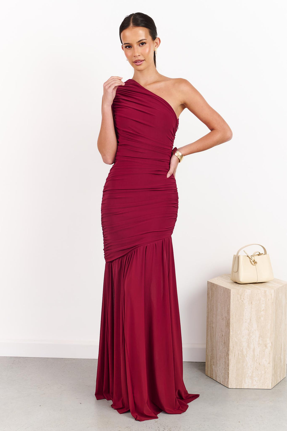 A woman wearing the ELLIATT Florencea one shoulder event gown in Wine Red, featuring a ruched bodice and draped maxi length skirt. She is standing in a front of a white wall.