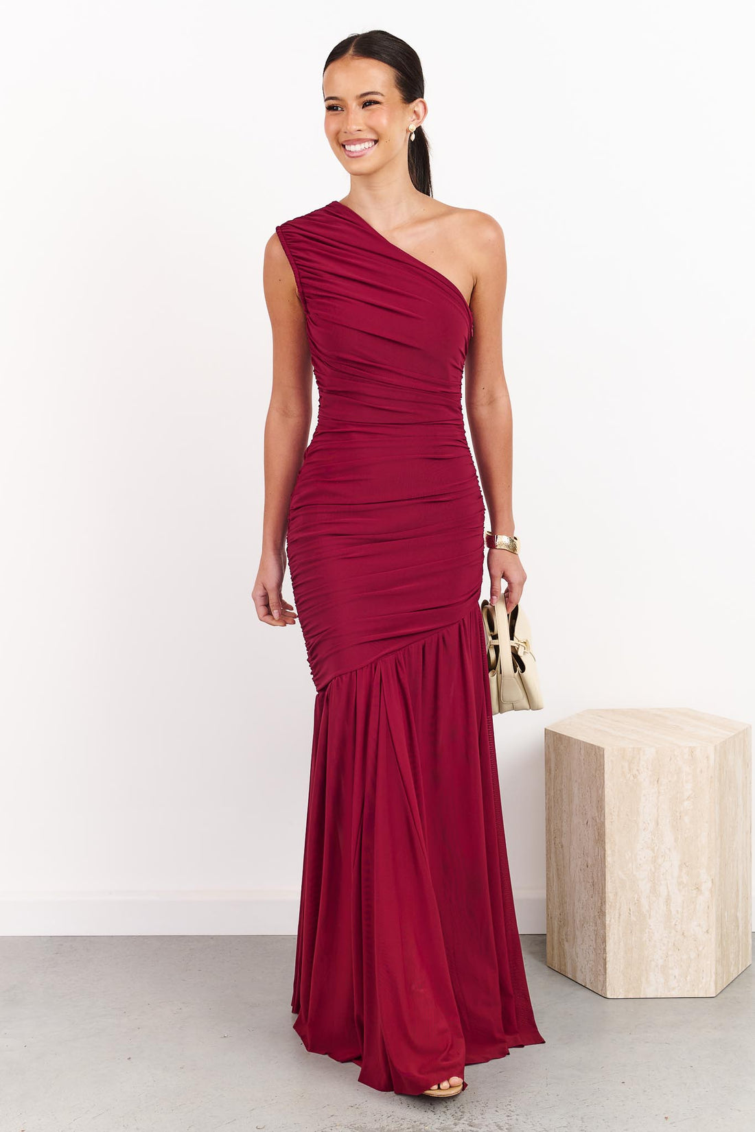 A woman wearing the ELLIATT Florencea one shoulder event gown in Wine Red, featuring a ruched bodice and draped maxi length skirt. She is standing in a front of a white wall.