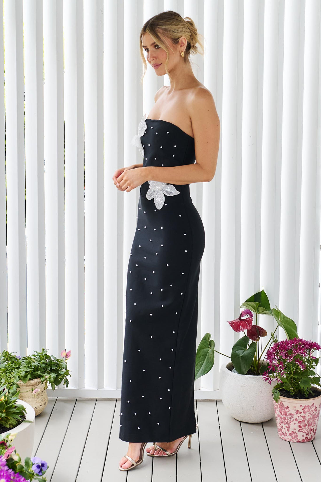 Side view woman modelling the ELLIATT Natasia embellished strapless black maxi dress with white floral appliqué flowers and pearl stud embellishments, photographed against a white panelled wall.