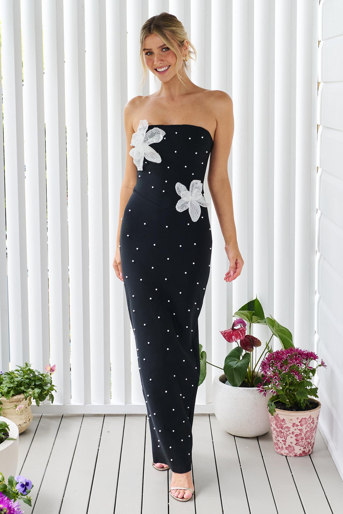 A woman modelling the ELLIATT Natasia embellished strapless black maxi dress with white floral appliqué flowers and pearl stud embellishments, photographed against a white panelled wall.