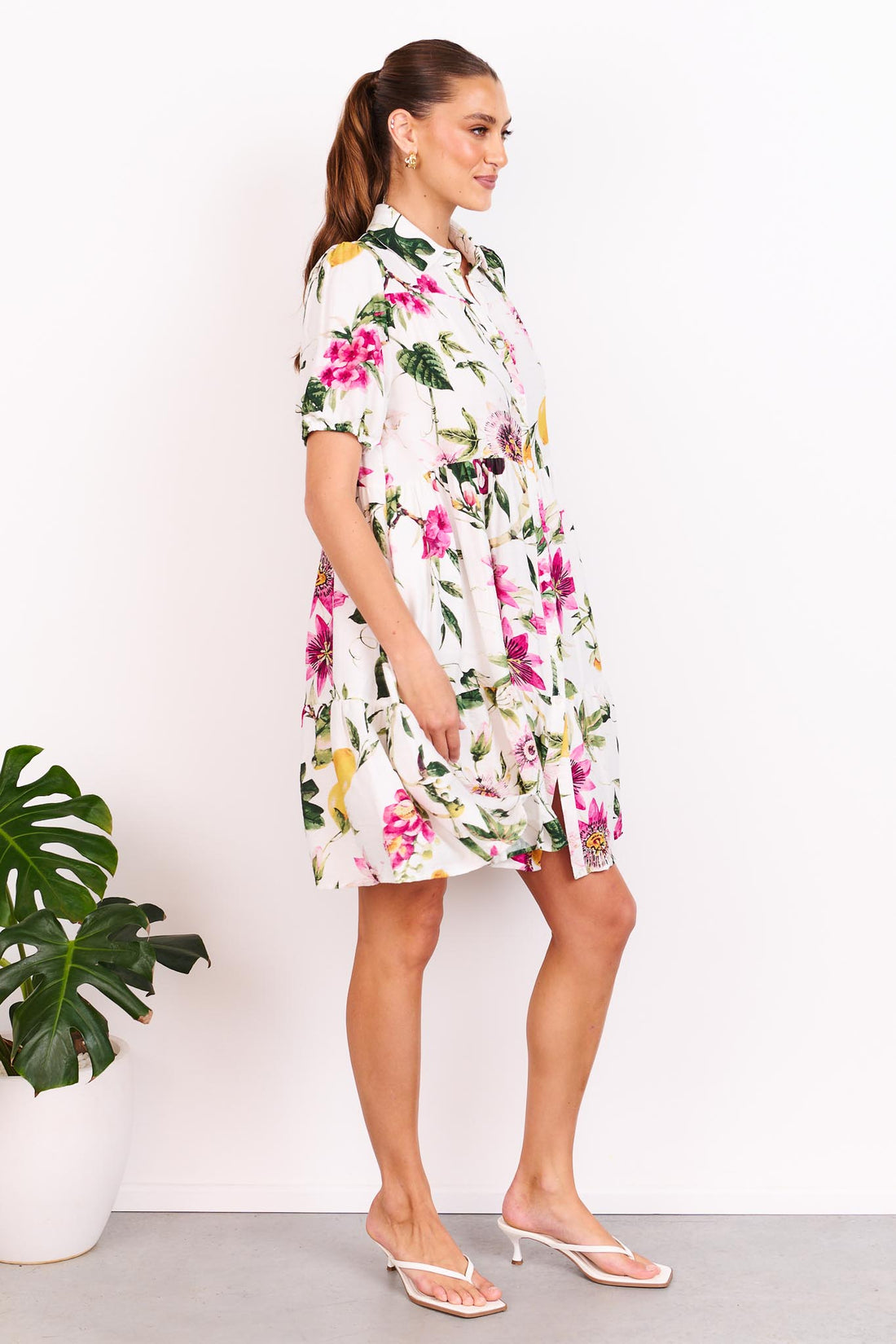 A woman wearing the minnie + MAXXIE Eden A-line shirt dress with short-sleeves, a collared neckline and a floral and lemon print, standing in front of a white wall.