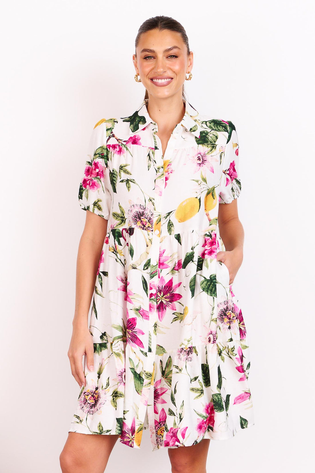 A woman wearing the minnie + MAXXIE Eden A-line shirt dress with short-sleeves, a collared neckline and a floral and lemon print, standing in front of a white wall.