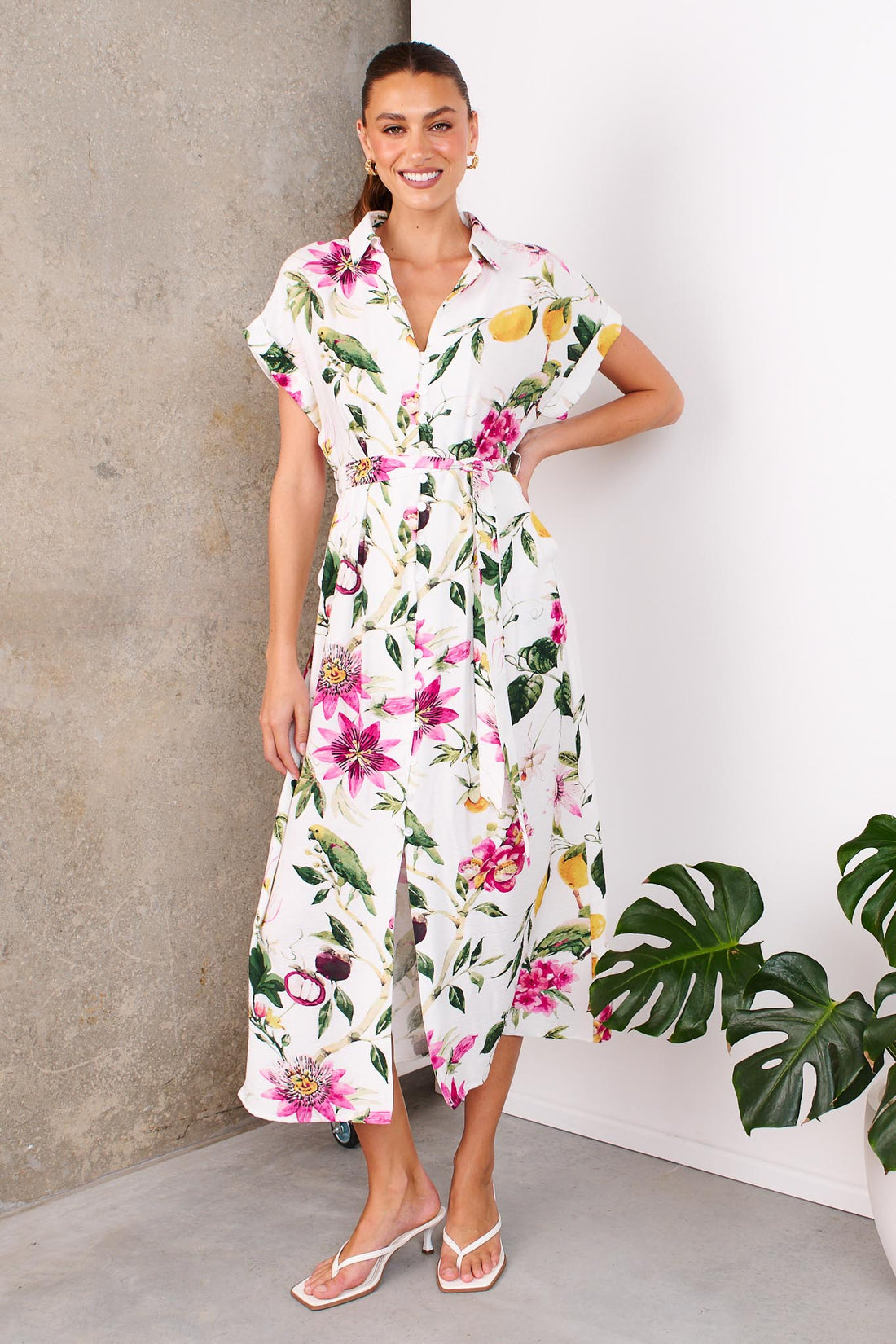 A woman wearing the minnie + MAXXIE Eden midi dress with a collared neckline, short sleeves, a botanical print and matching waist tie, standing in front of a white and concrete wall.