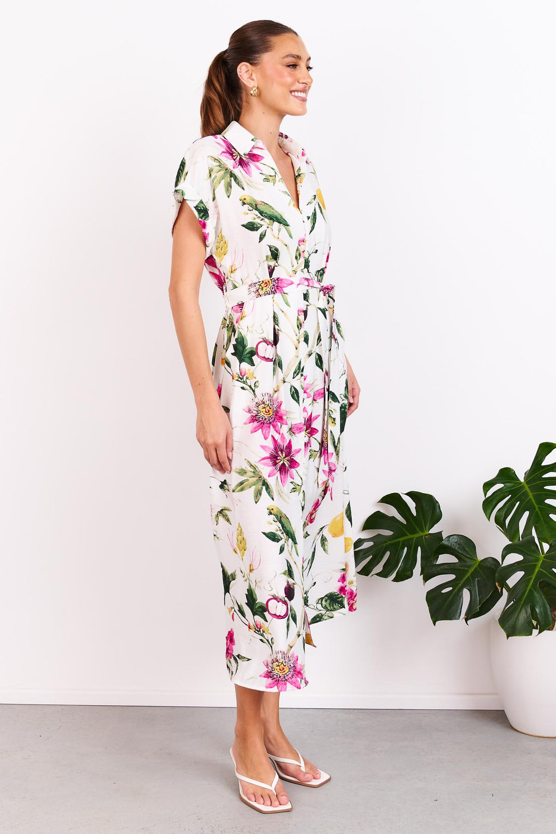 A woman wearing the minnie + MAXXIE Eden midi dress with a collared neckline, short sleeves, a botanical print and matching waist tie, standing in front of a white wall.