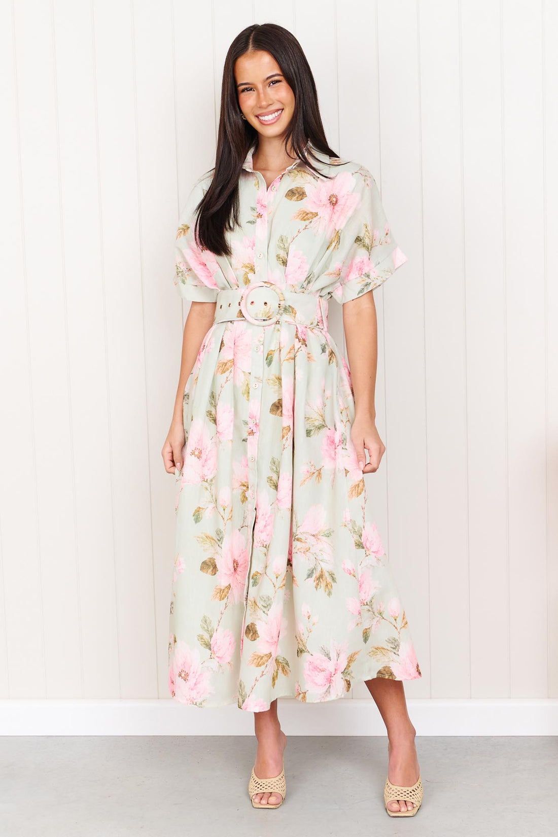 A woman wearing the minnie + MAXXIE Elodie Midi Dress in soft sage green with pink flowers, featuring short sleeves and a belted waist.