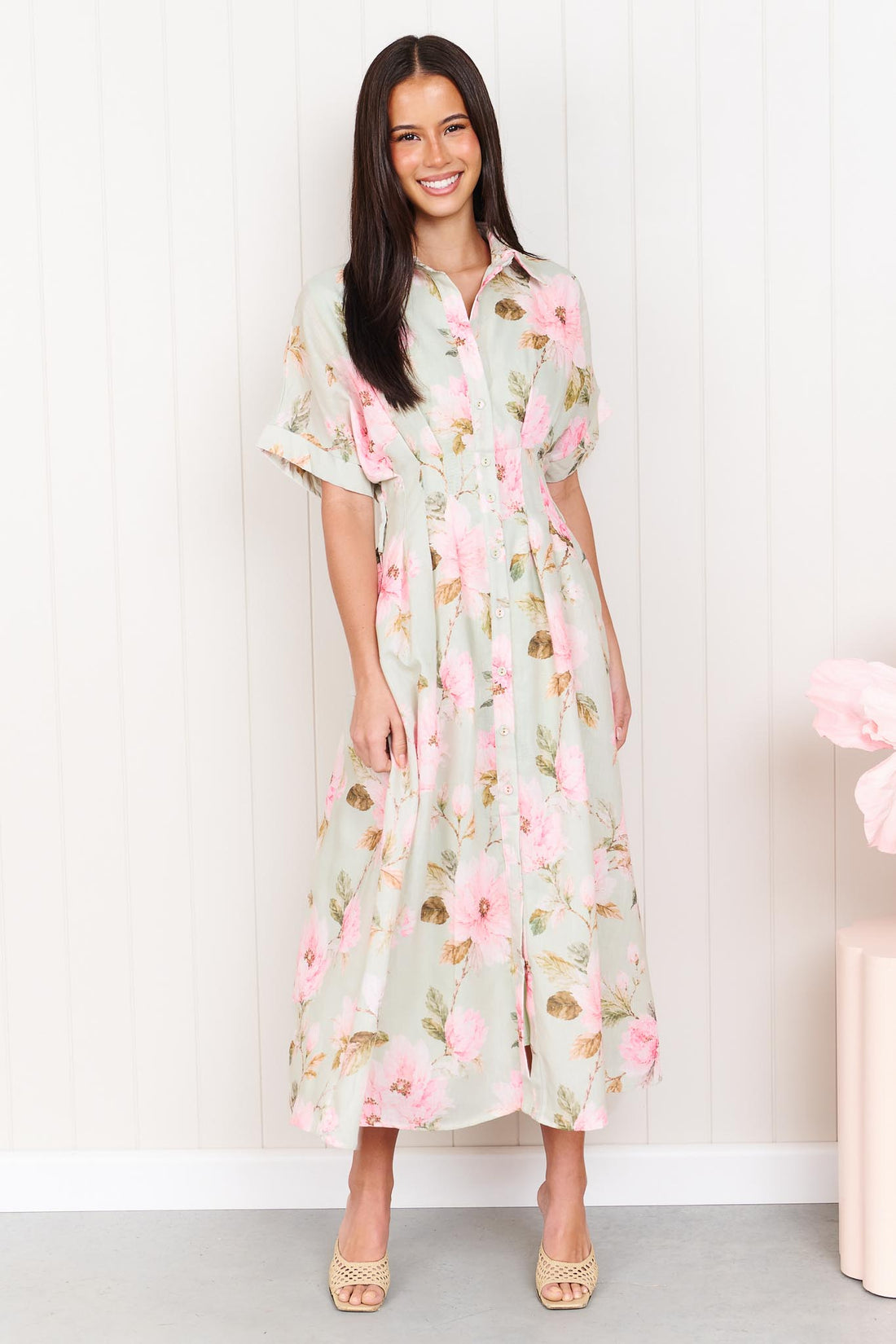 A woman wearing the minnie + MAXXIE Elodie Midi Dress in soft sage green with pink flowers, featuring short sleeves and a pleated waist.