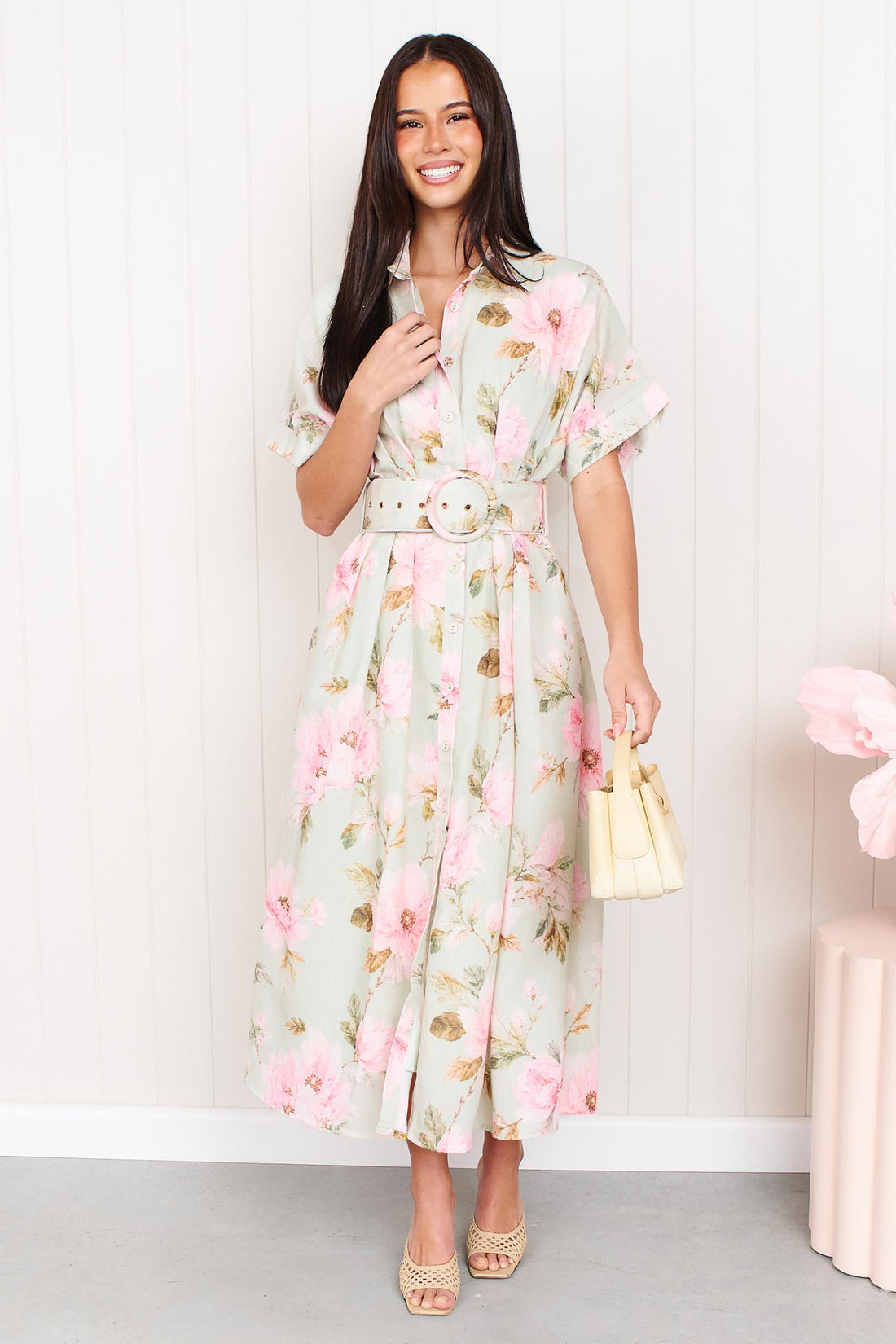 A woman wearing the minnie + MAXXIE Elodie Midi Dress in soft sage green with pink flowers, featuring short sleeves and a belted waist.