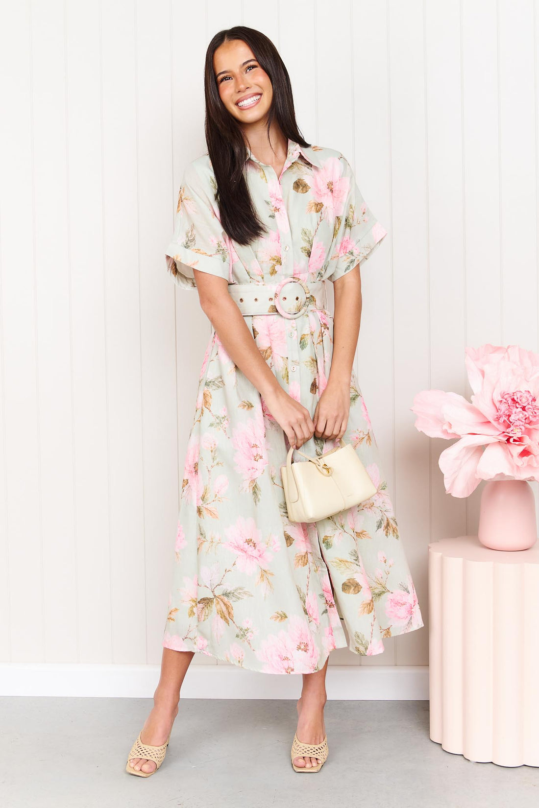 A woman wearing the minnie + MAXXIE Elodie Midi Dress in soft sage green with pink flowers, featuring short sleeves and a belted waist.