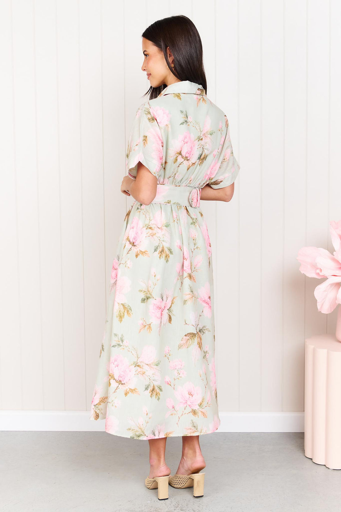 Back view of a woman wearing the minnie + MAXXIE Elodie Midi Dress in soft sage green with pink flowers, featuring short sleeves and a belted waist.