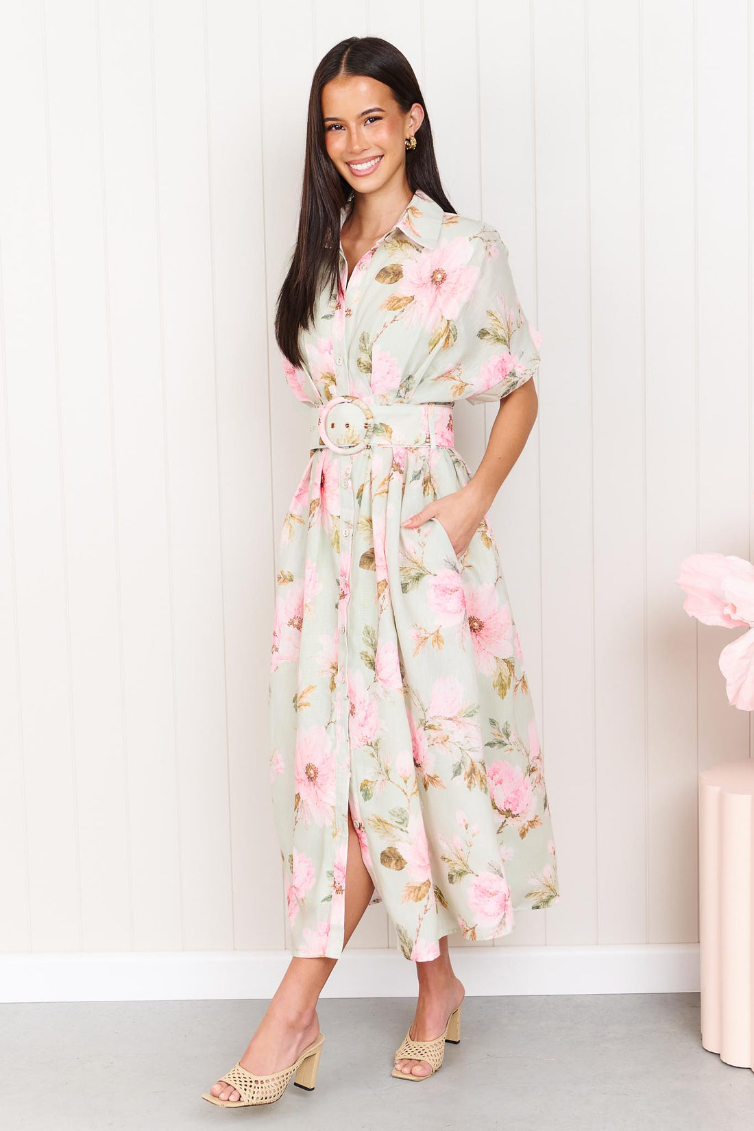 A woman wearing the minnie + MAXXIE Elodie Midi Dress in soft sage green with pink flowers, featuring short sleeves, a belted waist and side pockets.