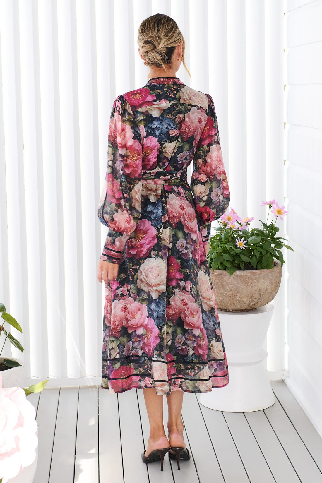Back view of a woman wearing the minnie + MAXXIE Fiora long-sleeve floral print midi dress with tie waist, standing in front of a white panelled wall. The dress features a multicolor floral pattern with pink, red, blue, and green tones on a black background.