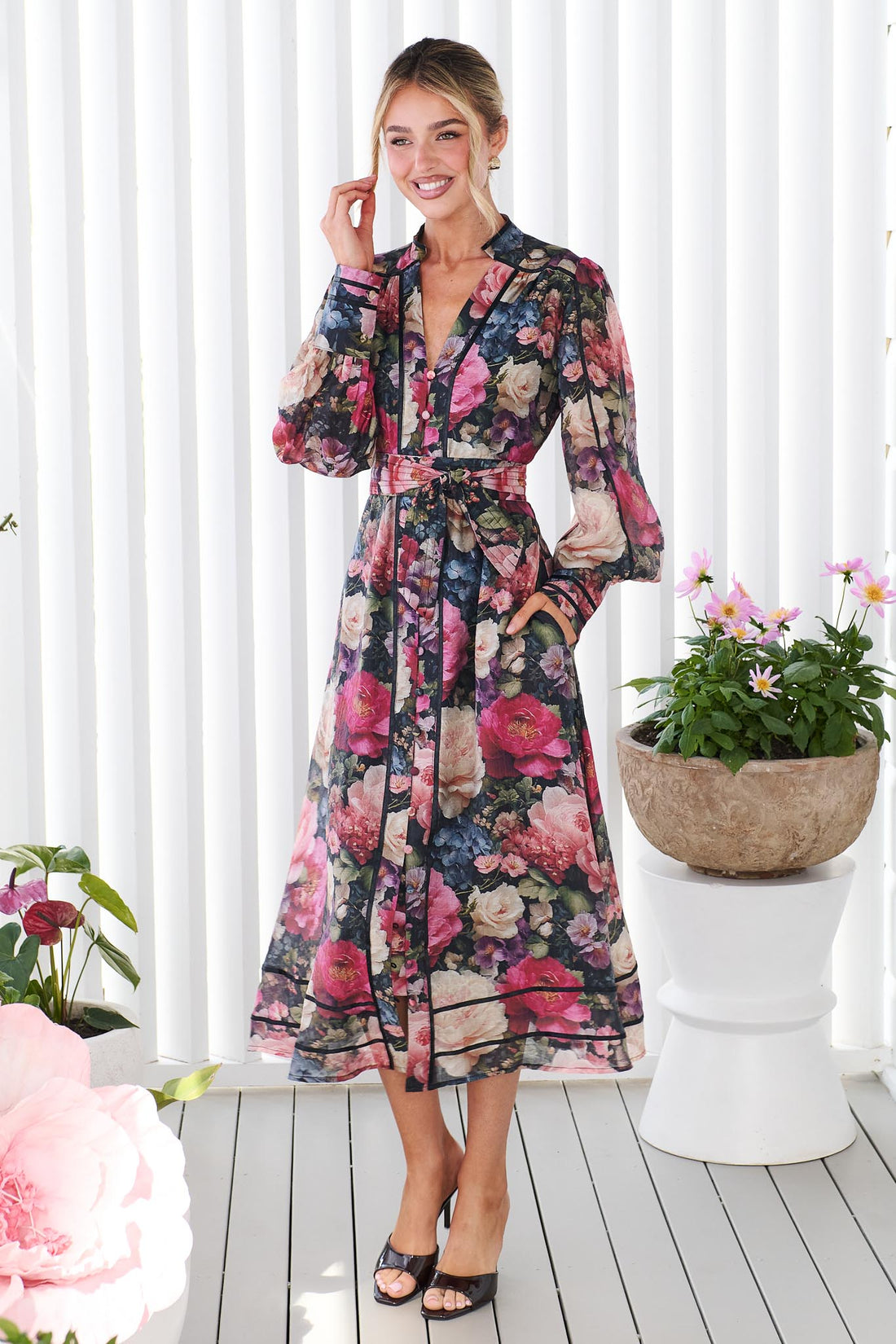 A woman wearing the minnie + MAXXIE Fiora long-sleeve floral print midi dress with tie waist, standing in front of a white panelled wall. The dress features a multicolor floral pattern with pink, red, blue, and green tones on a black background.