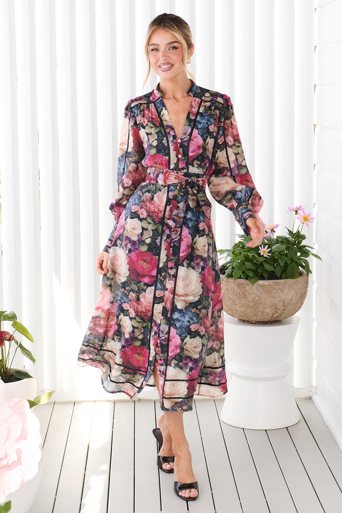 A woman wearing the minnie + MAXXIE Fiora long-sleeve floral print midi dress with tie waist, standing in front of a white panelled wall. The dress features a multicolor floral pattern with pink, red, blue, and green tones on a black background.