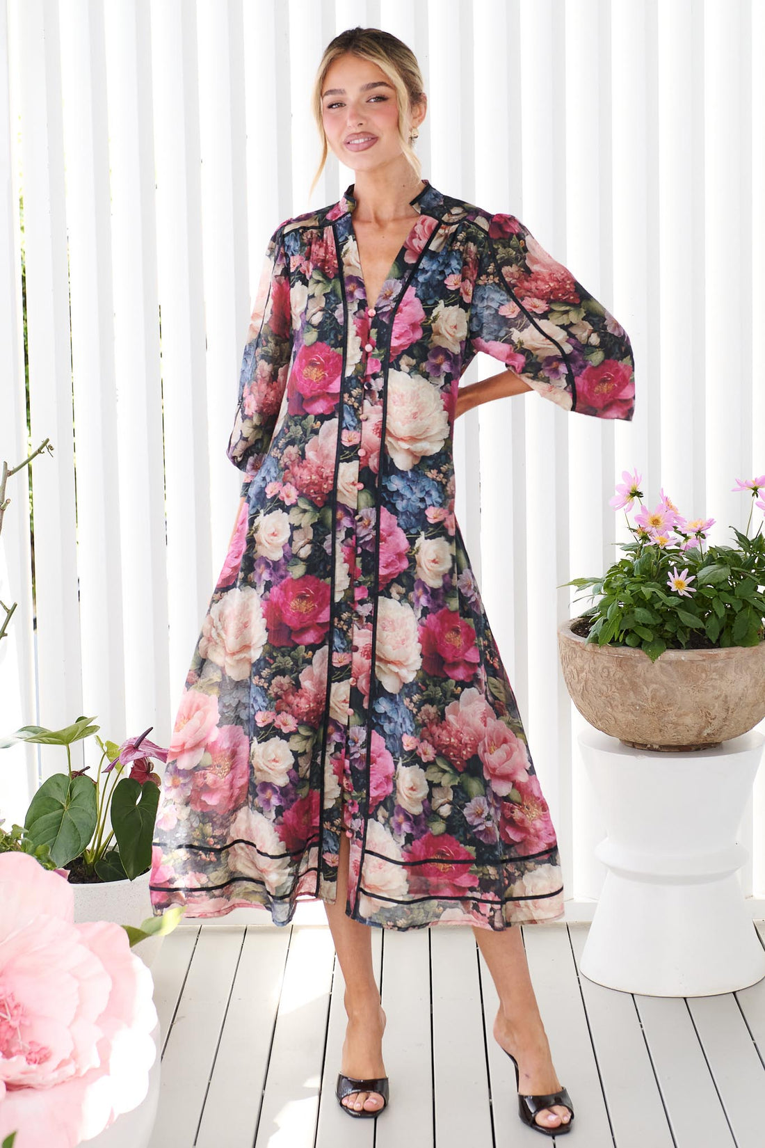 A woman wearing the minnie + MAXXIE Fiora long-sleeve floral print midi dress, standing in front of a white panelled wall. The dress features a multicolor floral pattern with pink, red, blue, and green tones on a black background.