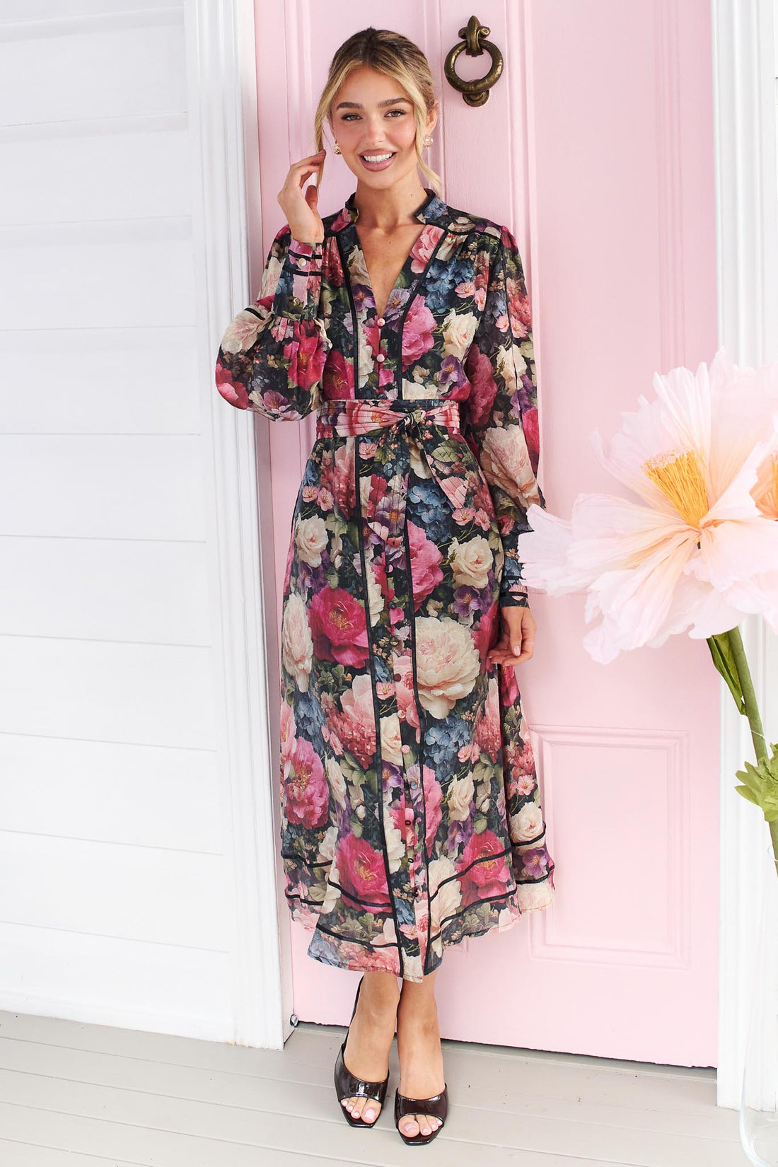 A woman wearing the minnie + MAXXIE Fiora long-sleeve floral print midi dress with tie waist, standing in front of a pink door.