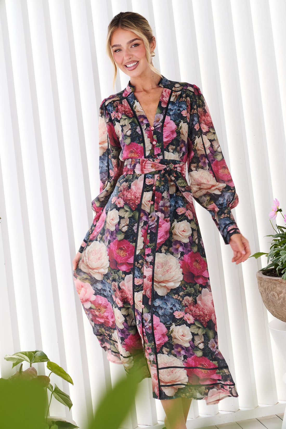 A woman wearing the minnie + MAXXIE Fiora long-sleeve floral print midi dress with tie waist, standing in front of a white panelled wall. The dress features a multicolor floral pattern with pink, red, blue, and green tones on a black background.