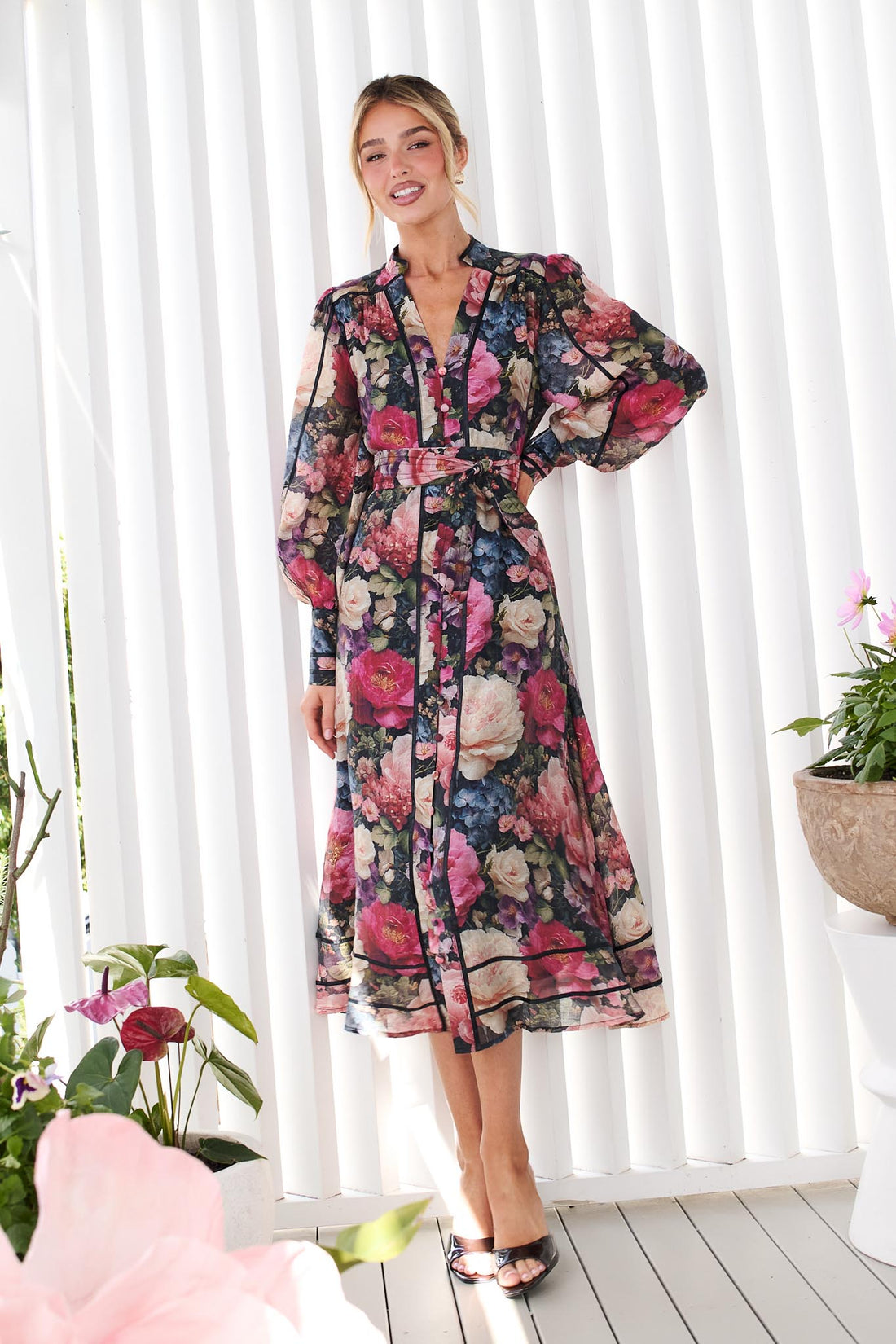 A woman wearing the minnie + MAXXIE Fiora long-sleeve floral print midi dress with tie waist, standing in front of a white panelled wall. The dress features a multicolor floral pattern with pink, red, blue, and green tones on a black background.