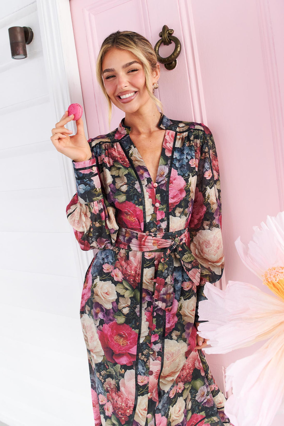A woman wearing the minnie + MAXXIE Fiora long-sleeve floral print midi dress with tie waist, standing in front of a pink door, holding a pink macaron. The dress features a multicolor floral pattern with pink, red, blue, and green tones on a black background.