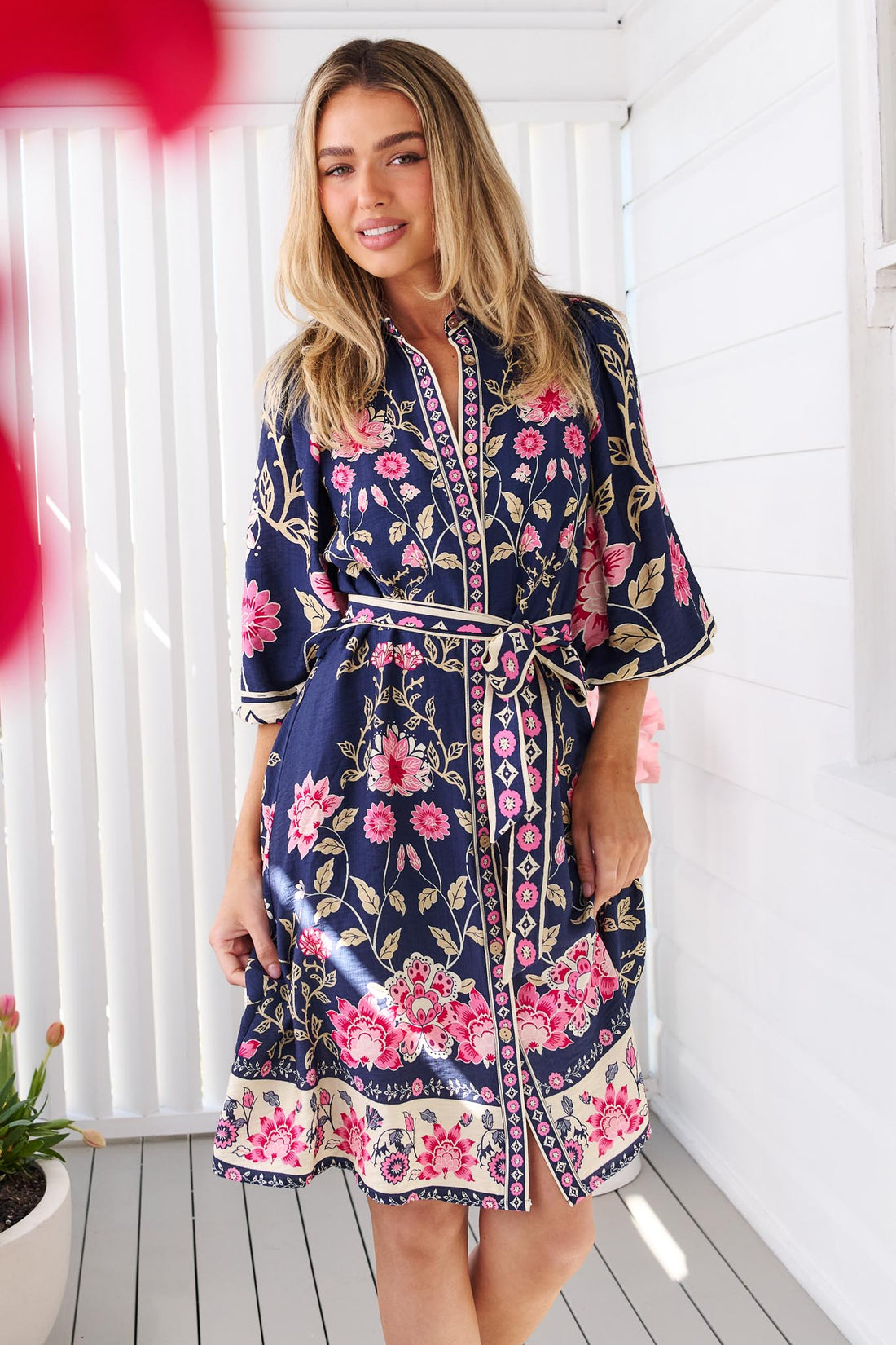 A woman wearing the minnie + MAXXIE Francesca dress in navy blue with a pink and green floral print, featuring a tie waist and billow sleeves, set against a bright white background.