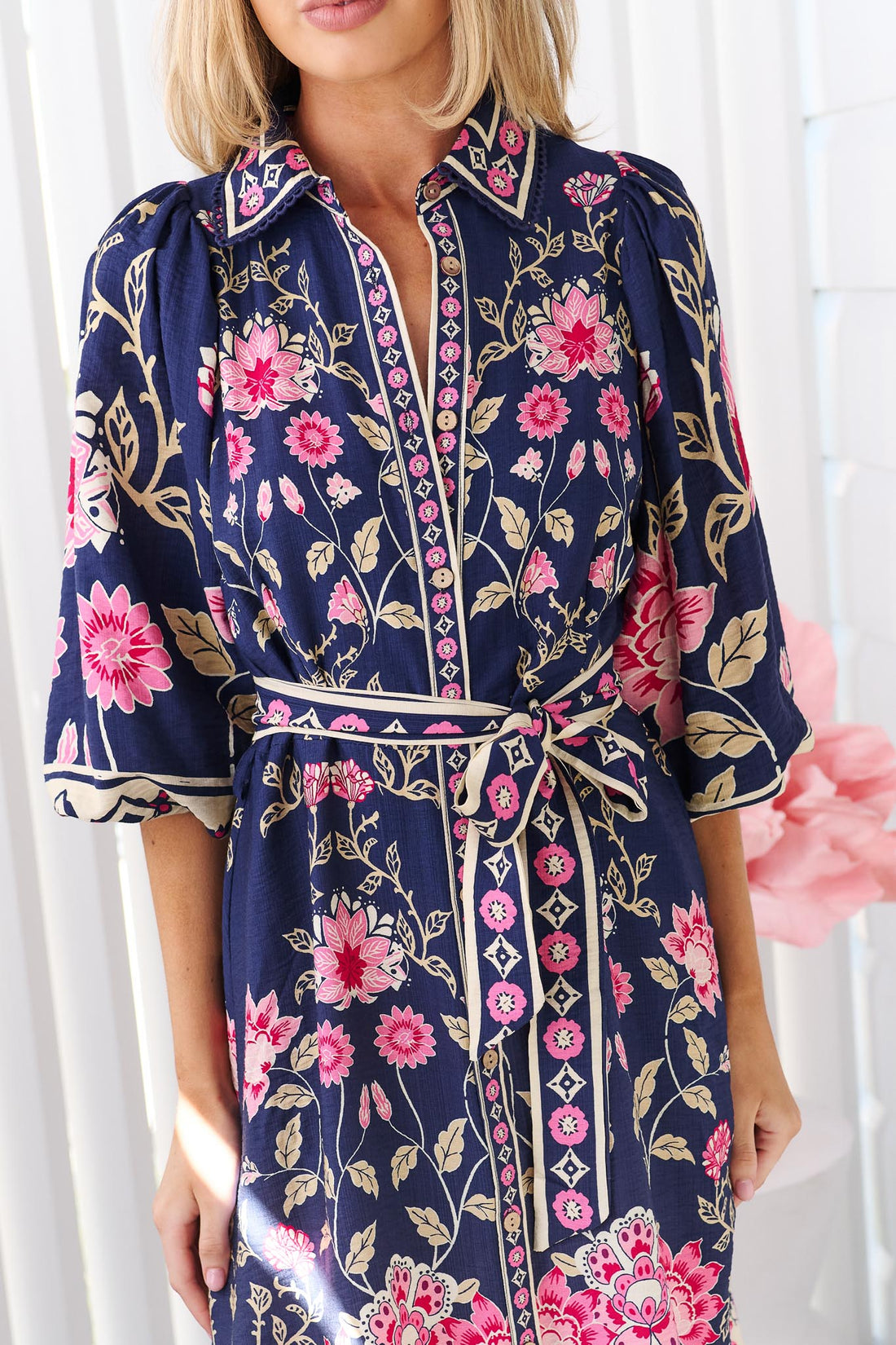 Close up of a woman wearing the minnie + MAXXIE Francesca dress in navy blue with a pink and green floral print, featuring a tie waist and 3/4 billow sleeves.