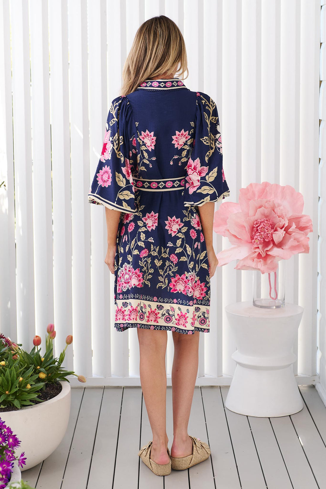 Back view of a woman wearing the minnie + MAXXIE Francesca dress in navy blue with a pink and green floral print, featuring a tie waist and billow sleeves, set against a bright white background.