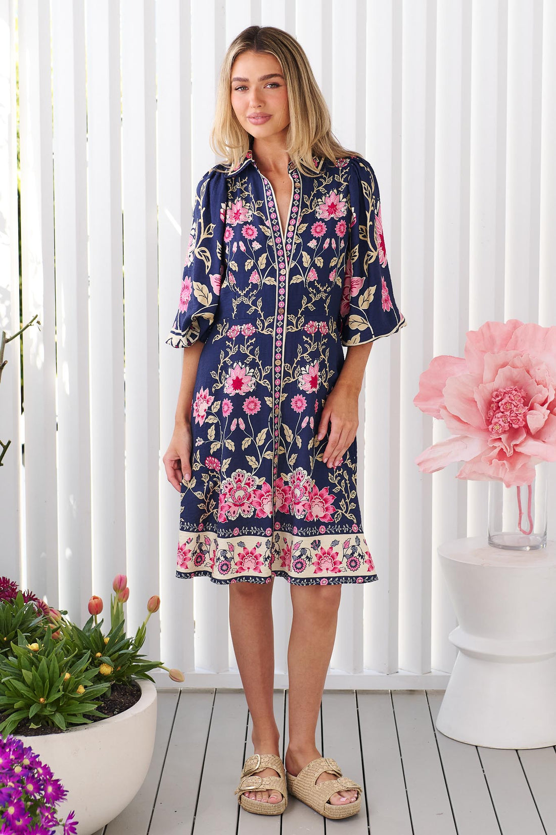 A woman wearing the minnie + MAXXIE Francesca dress in navy blue with a pink and green floral print and billow sleeves, set against a bright white background.