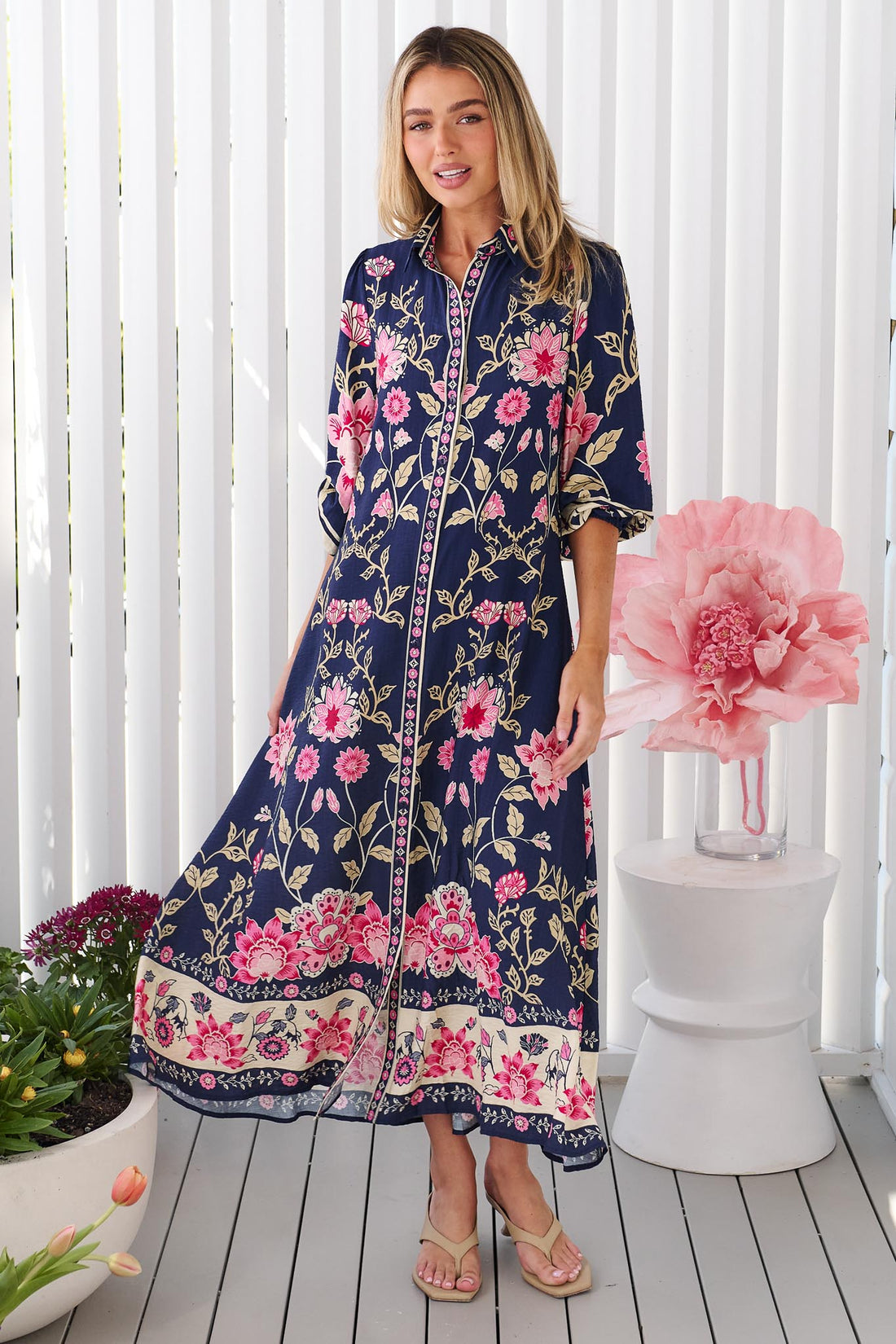 A woman wearing the minnie + MAXXIE Francesca Midi Dress in navy blue with a pink and beige floral print, standing in front of a white panelled wall.