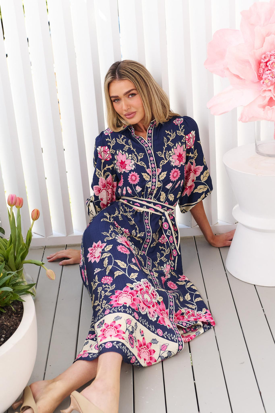 A woman sitting on a white deck wearing the minnie + MAXXIE Francesca Midi Dress in navy blue with a pink and beige floral print and matching waist tie.
