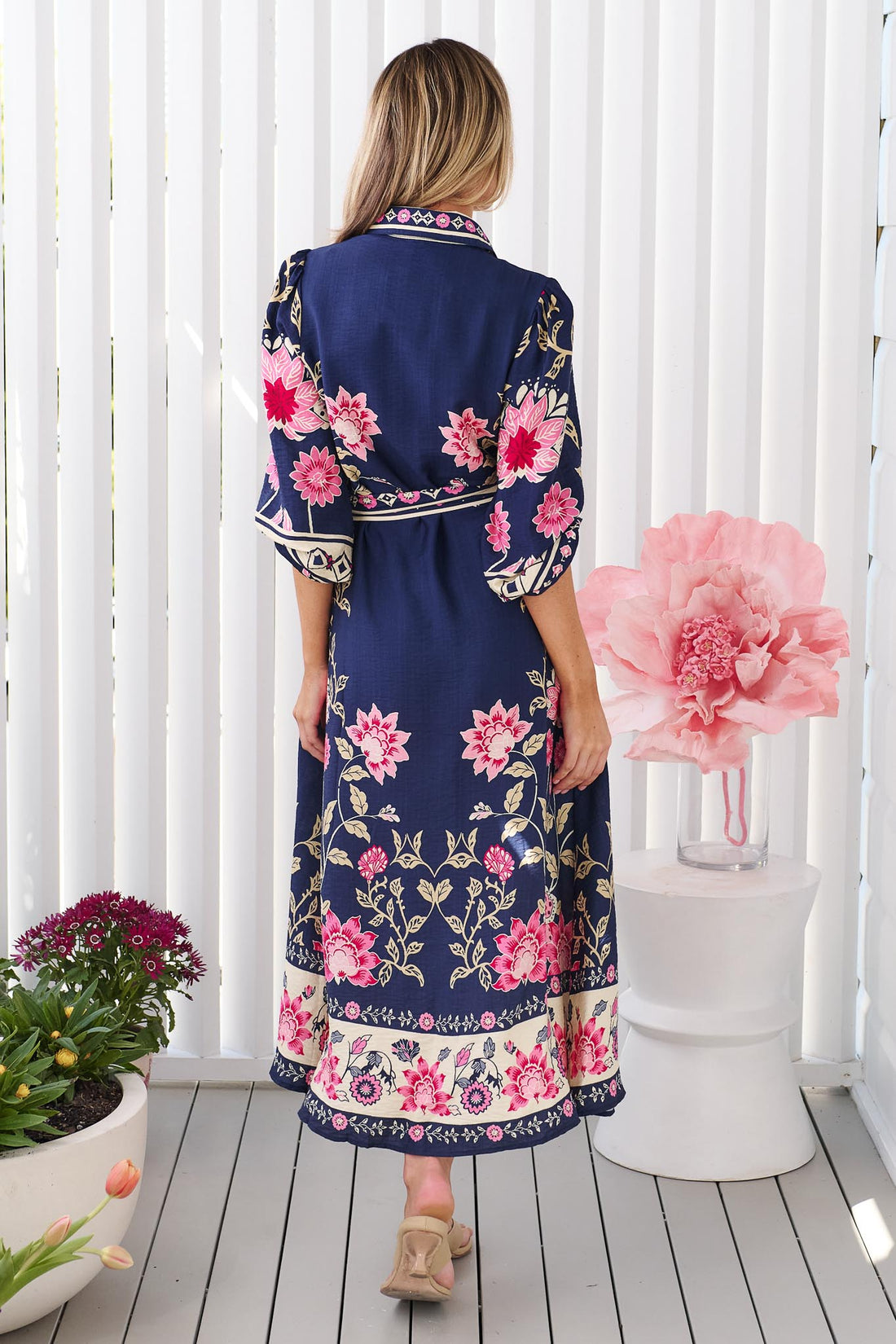 Back view of a woman wearing the minnie + MAXXIE Francesca Midi Dress in navy blue with a pink and beige floral print and matching waist tie, standing in front of a white panelled wall.
