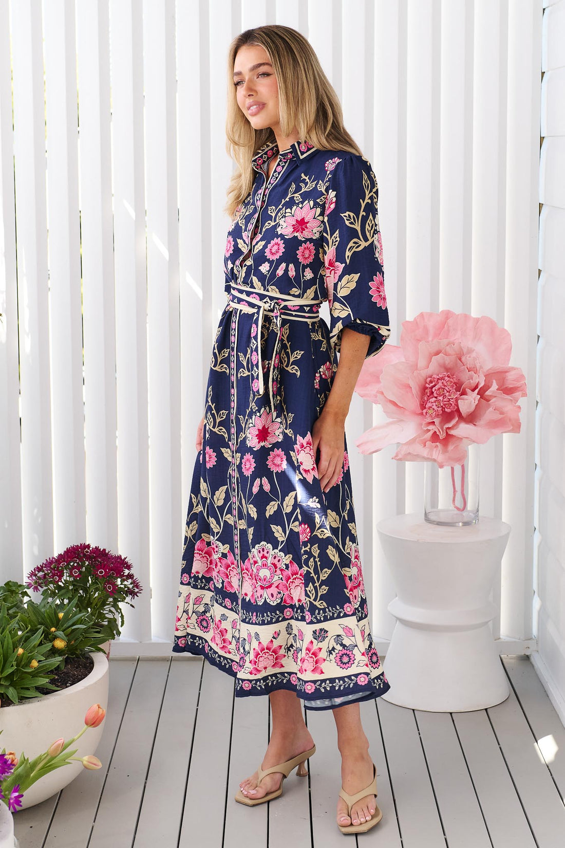 Side view of a woman wearing the minnie + MAXXIE Francesca Midi Dress in navy blue with a pink and beige floral print and matching waist tie, standing in front of a white panelled wall.