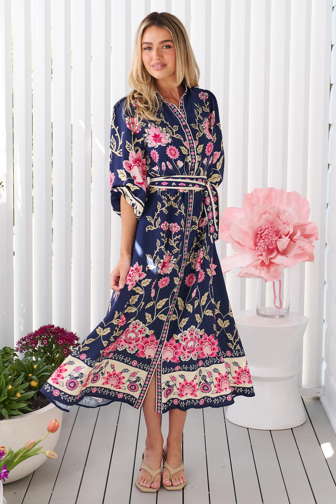 A woman wearing the minnie + MAXXIE Francesca Midi Dress in navy blue with a pink and beige floral print and matching waist tie, standing in front of a white panelled wall.