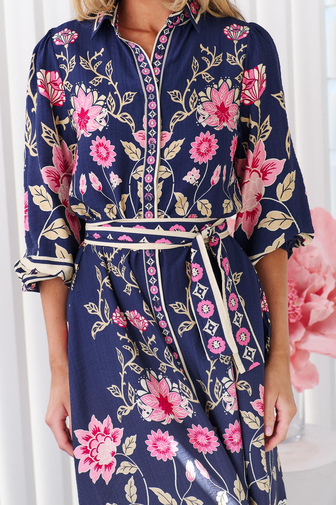 Close up of a woman wearing the minnie + MAXXIE Francesca Midi Dress in navy blue with a pink and beige floral print and matching waist tie, standing in front of a white panelled wall.